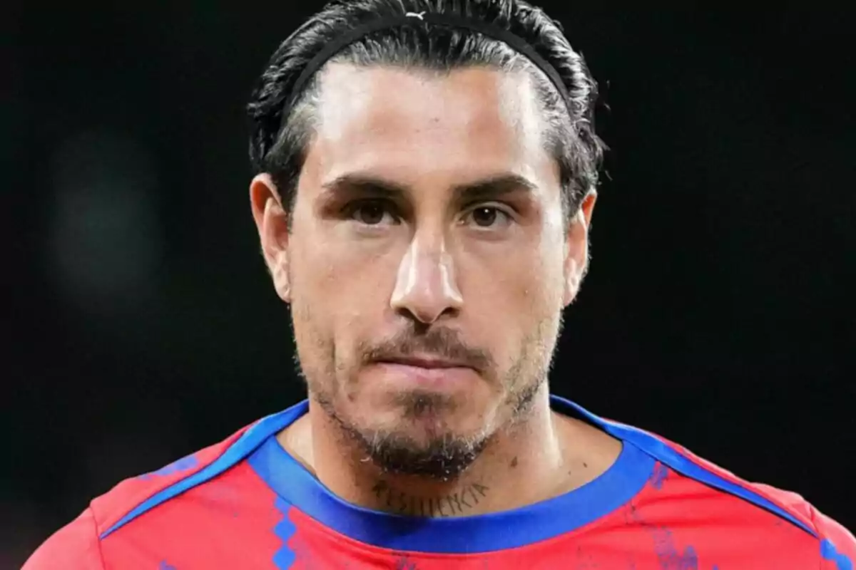 Un hombre con cabello oscuro y una cinta en la cabeza lleva una camiseta roja con detalles azules y tiene un tatuaje en el cuello.