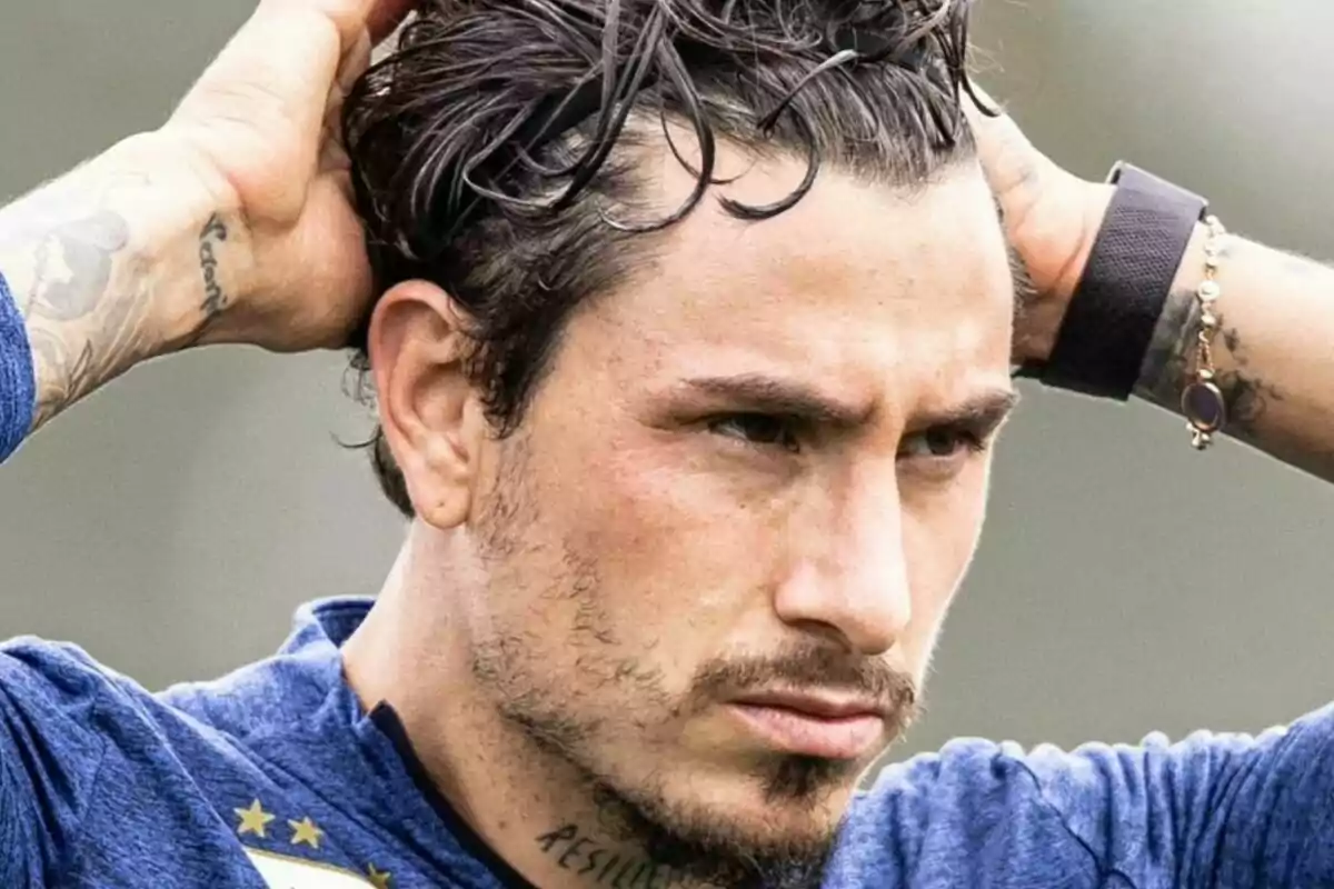 Hombre con expresión seria acomodándose el cabello con ambas manos, viste camiseta azul y tiene tatuajes visibles en el cuello y brazo