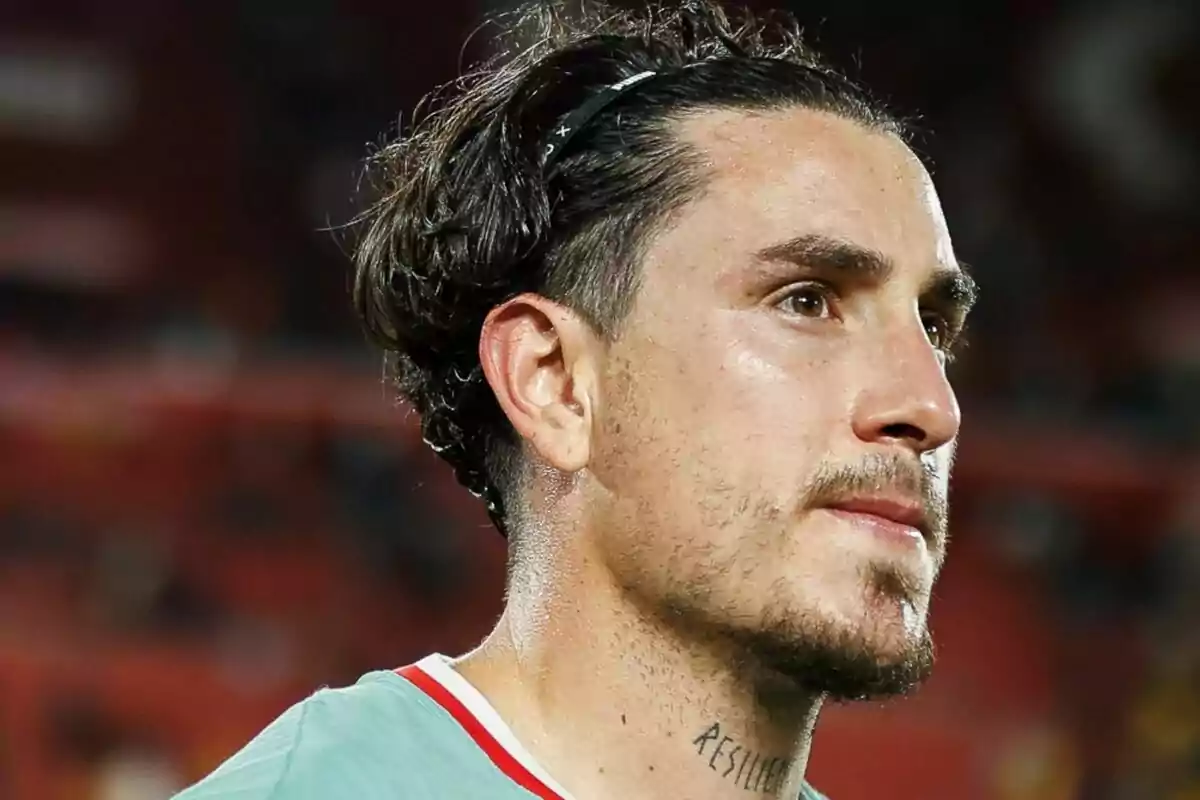 Un hombre con cabello oscuro y barba ligera mirando hacia la derecha en un entorno deportivo.