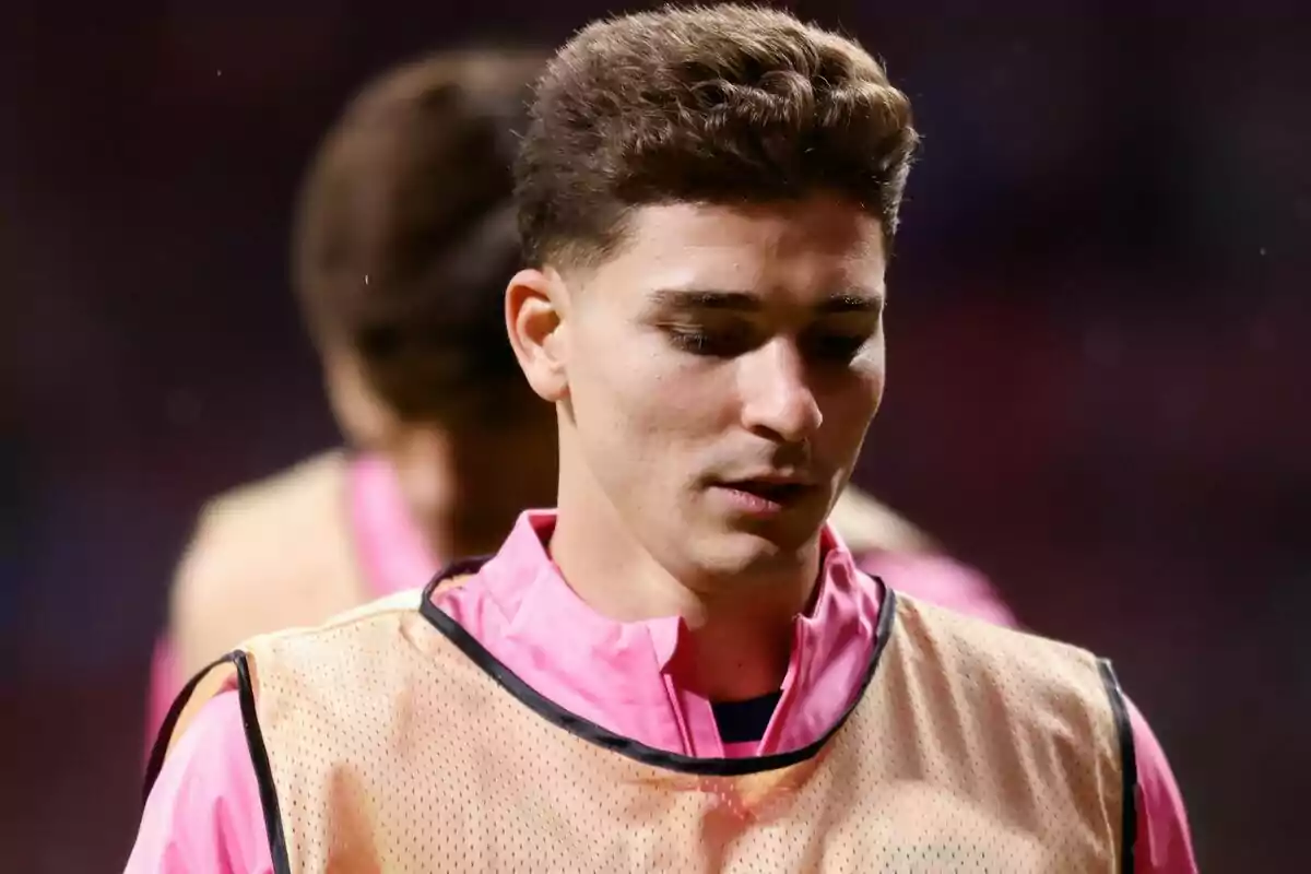 Un jugador de fútbol con uniforme rosa y chaleco de entrenamiento está mirando hacia abajo en un campo deportivo.