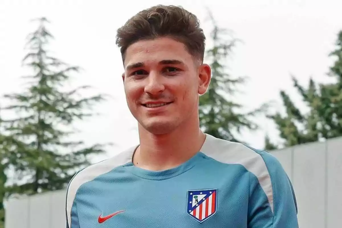 Un hombre con una camiseta de entrenamiento del Atlético de Madrid sonríe frente a un fondo de árboles.