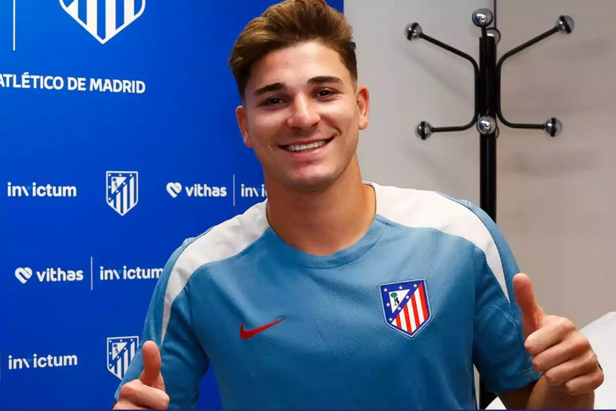 Un joven sonriente con una camiseta del Atlético de Madrid, posando frente a un fondo azul con logotipos del equipo y de patrocinadores.