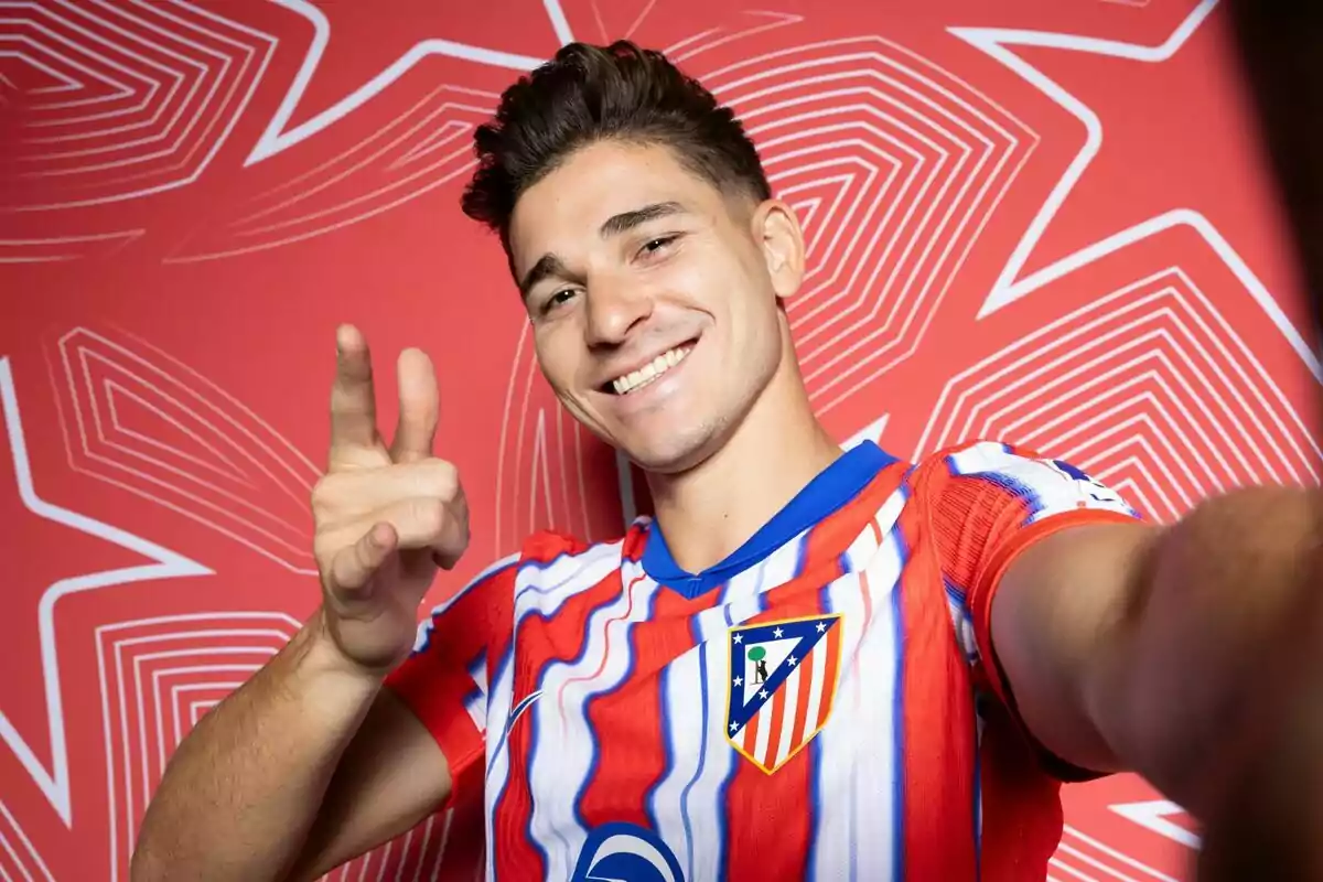 Un jugador de fútbol con la camiseta del Atlético de Madrid sonríe y hace un gesto con la mano mientras se toma una selfie frente a un fondo rojo con patrones geométricos.