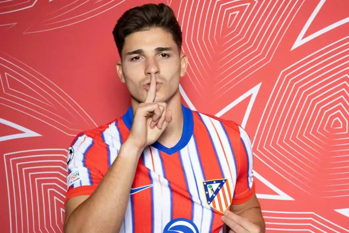 Un jugador de fútbol con la camiseta del Atlético de Madrid hace un gesto de silencio con el dedo índice sobre los labios frente a un fondo rojo con patrones geométricos.