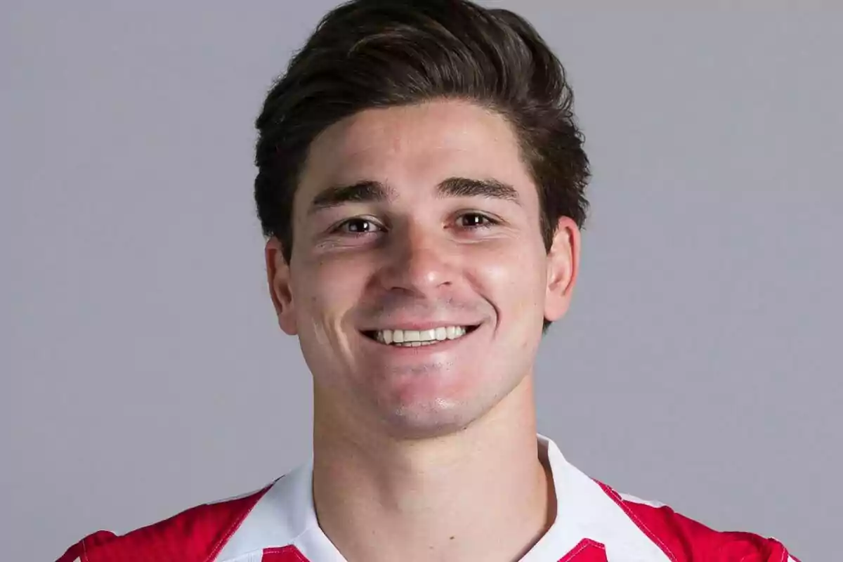 Joven con cabello castaño y corto sonriendo, vistiendo una camiseta deportiva roja con blanco sobre fondo gris.