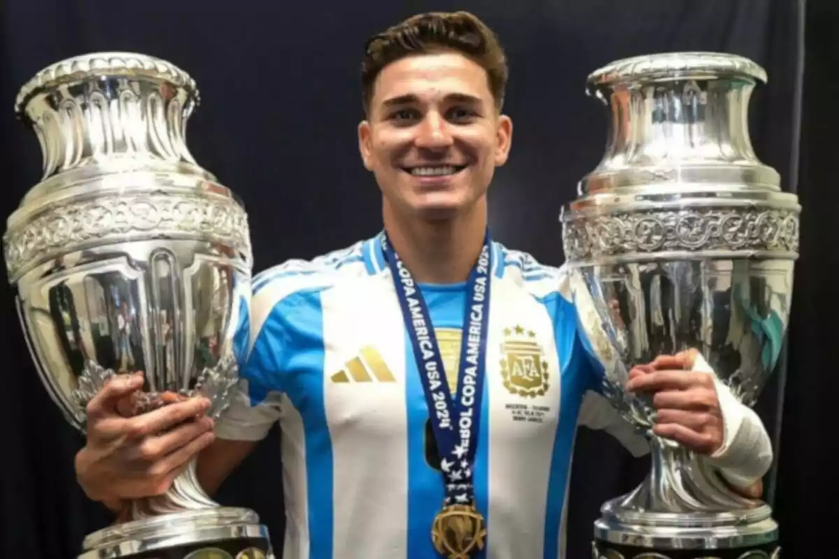 Un jugador de fútbol con la camiseta de la selección argentina sostiene dos trofeos grandes y plateados, mientras sonríe a la cámara.