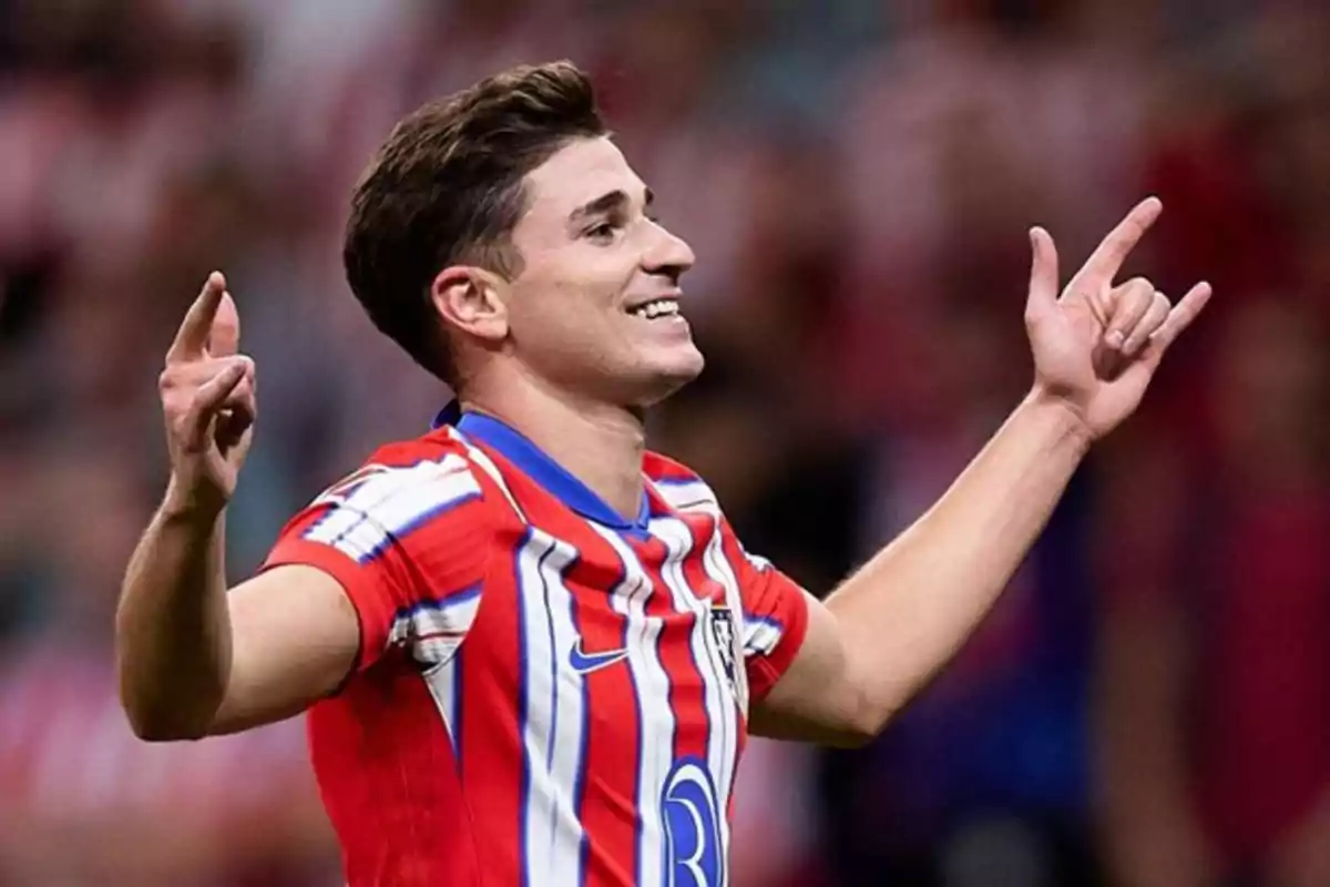 Un jugador de fútbol celebrando un gol con los brazos levantados y sonriendo, vistiendo una camiseta a rayas rojas, blancas y azules.