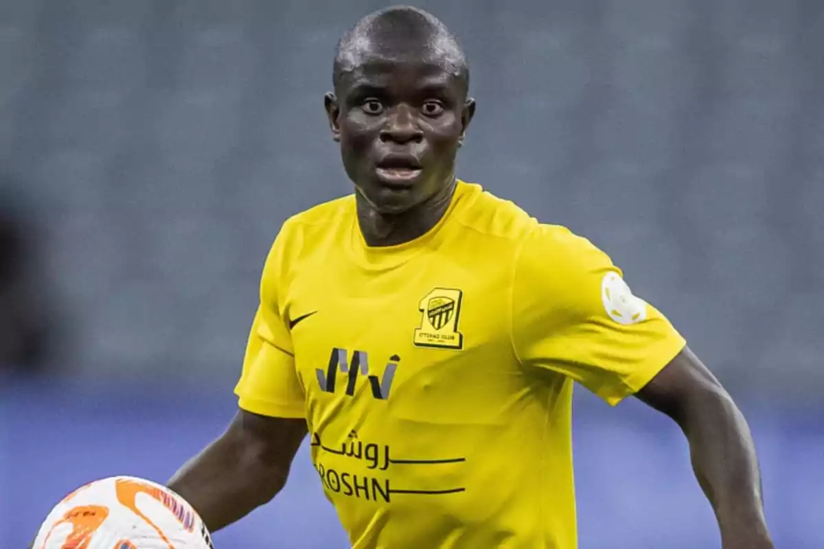 Jugador de fútbol con camiseta amarilla del Al-Ittihad Club sosteniendo un balón en el campo.