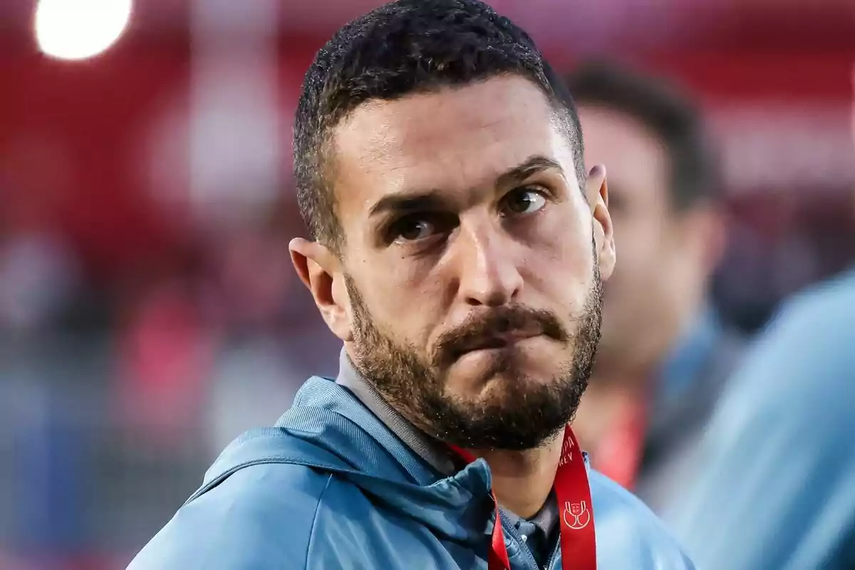 Un hombre con barba y cabello corto, vistiendo una chaqueta azul y una medalla roja, mira hacia un lado con expresión seria.