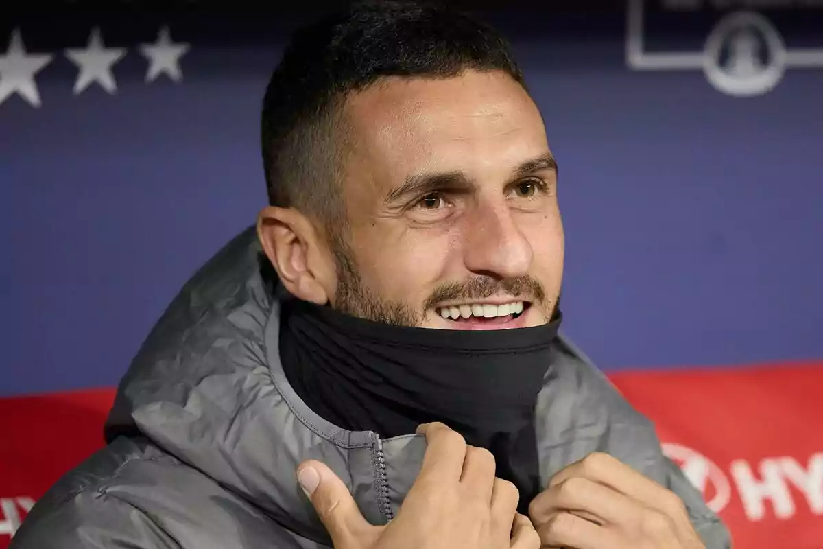 Un hombre sonriente con barba corta y chaqueta gris ajustándose una bufanda negra.