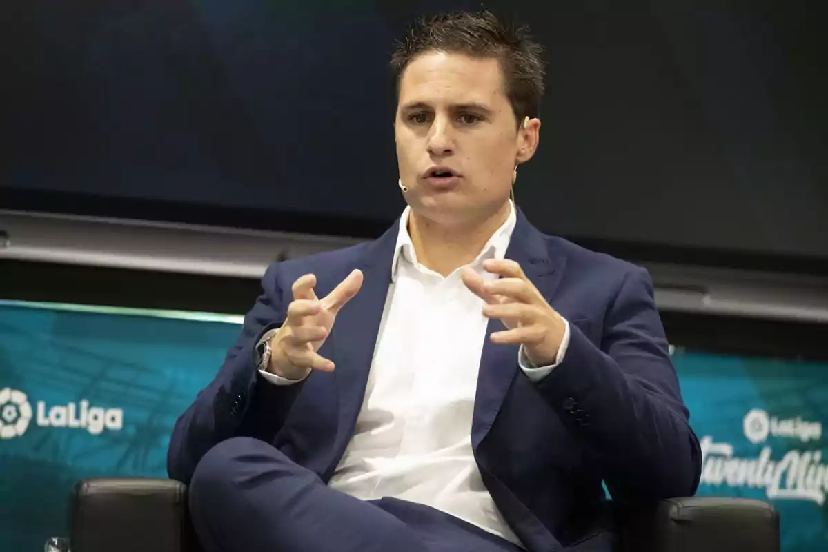 A man in a blue suit and white shirt is speaking while gesturing with his hands, sitting on a stage with the LaLiga logo in the background.