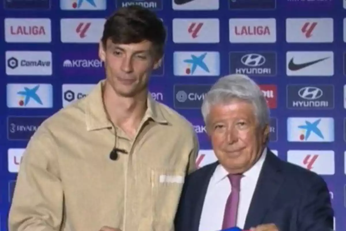 Dos hombres posan juntos frente a un fondo con logotipos de patrocinadores deportivos.