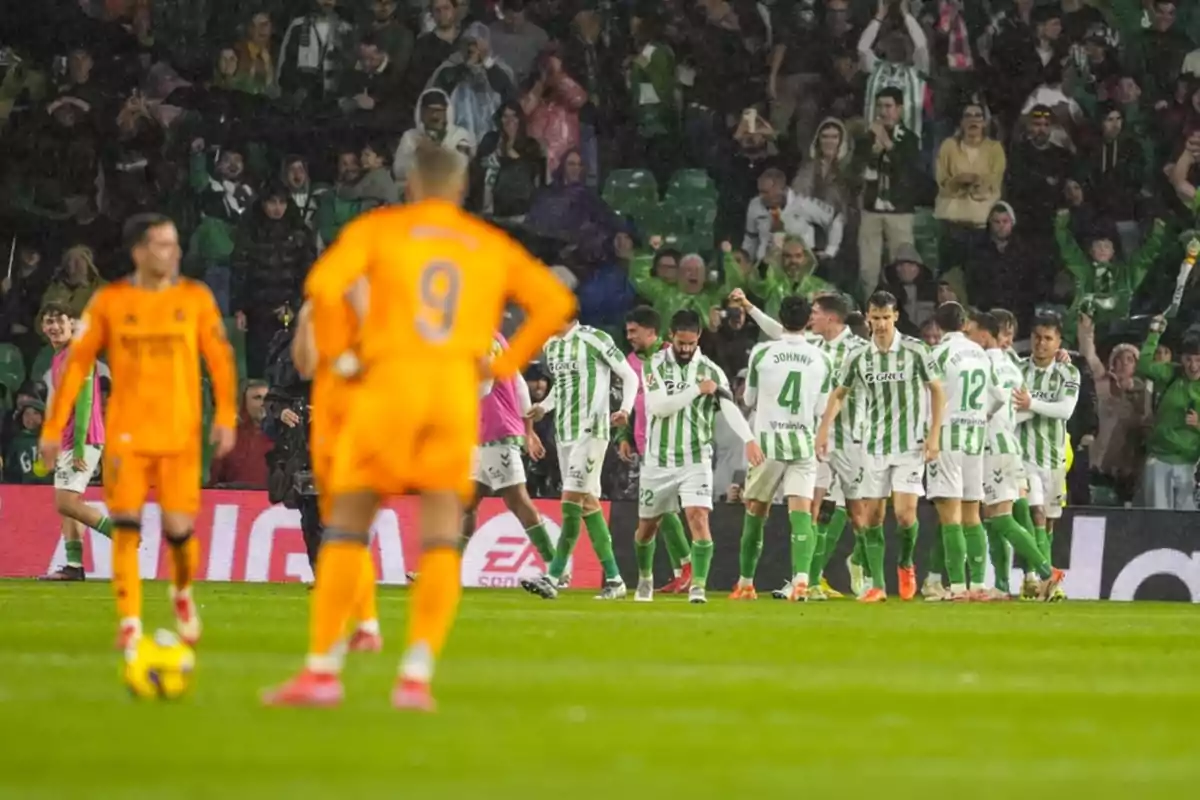 Jugadores de un equipo de fútbol con uniformes verdes y blancos celebran en el campo mientras los jugadores del equipo contrario, vestidos de naranja, observan.