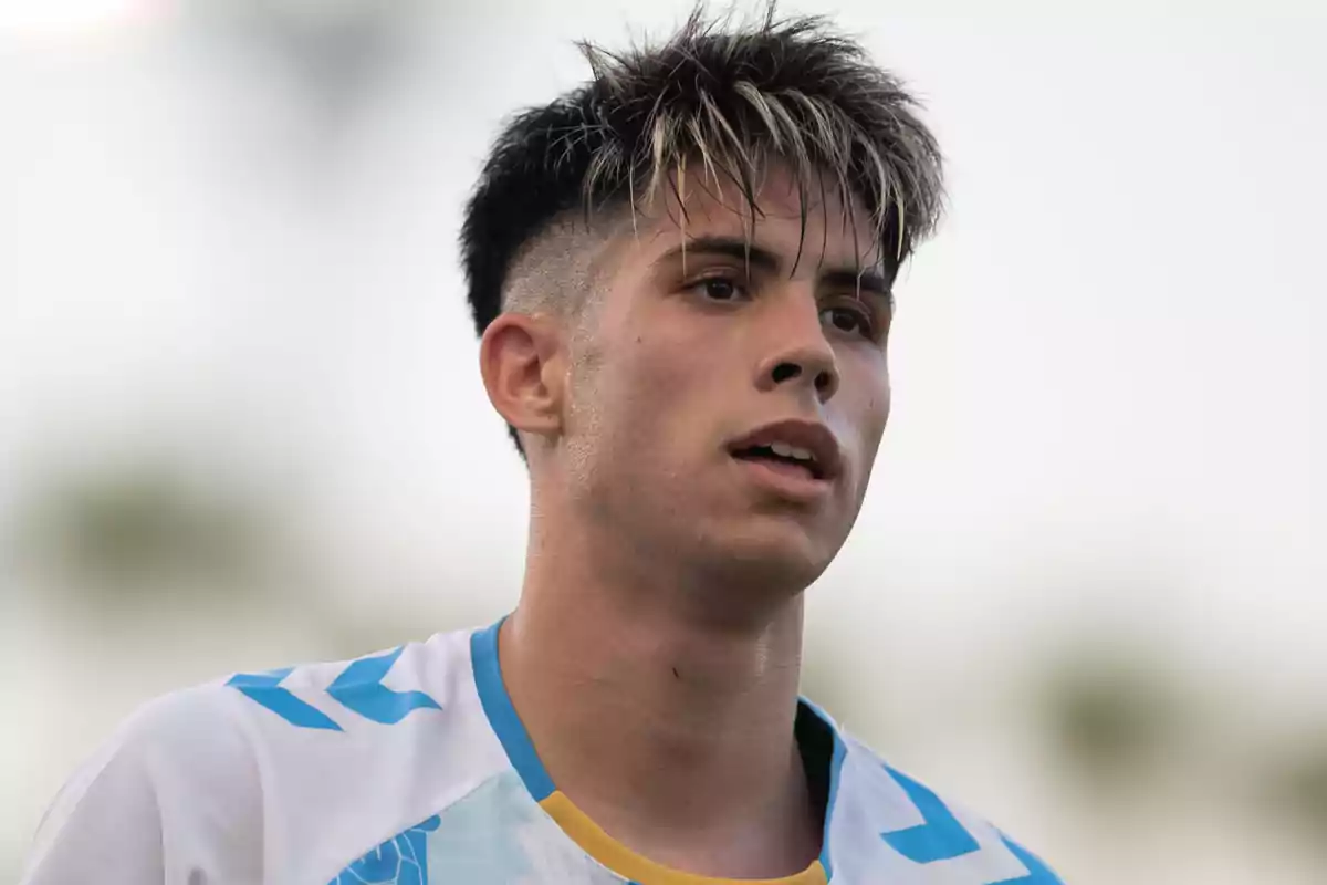 Un joven con camiseta deportiva blanca y detalles azules y amarillos mira hacia adelante con expresión seria.