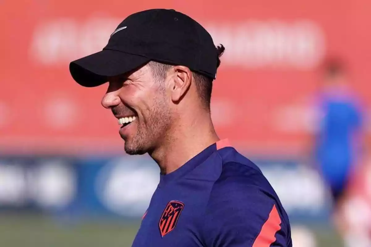 Hombre con gorra negra y camiseta deportiva sonríe en un campo de entrenamiento.