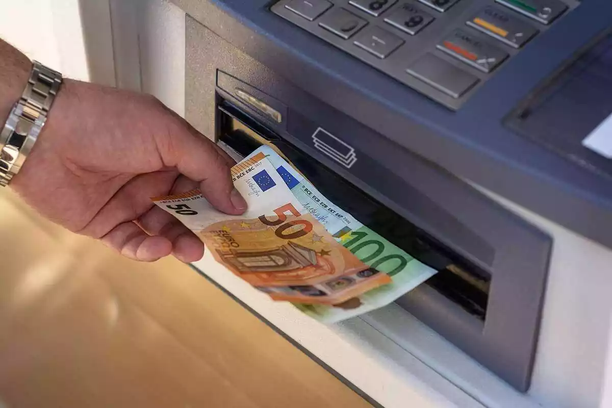 Mano izquierda con reloj metálico sacando un billete de 50 euros y un billete de 100 euros de un cajero automático
