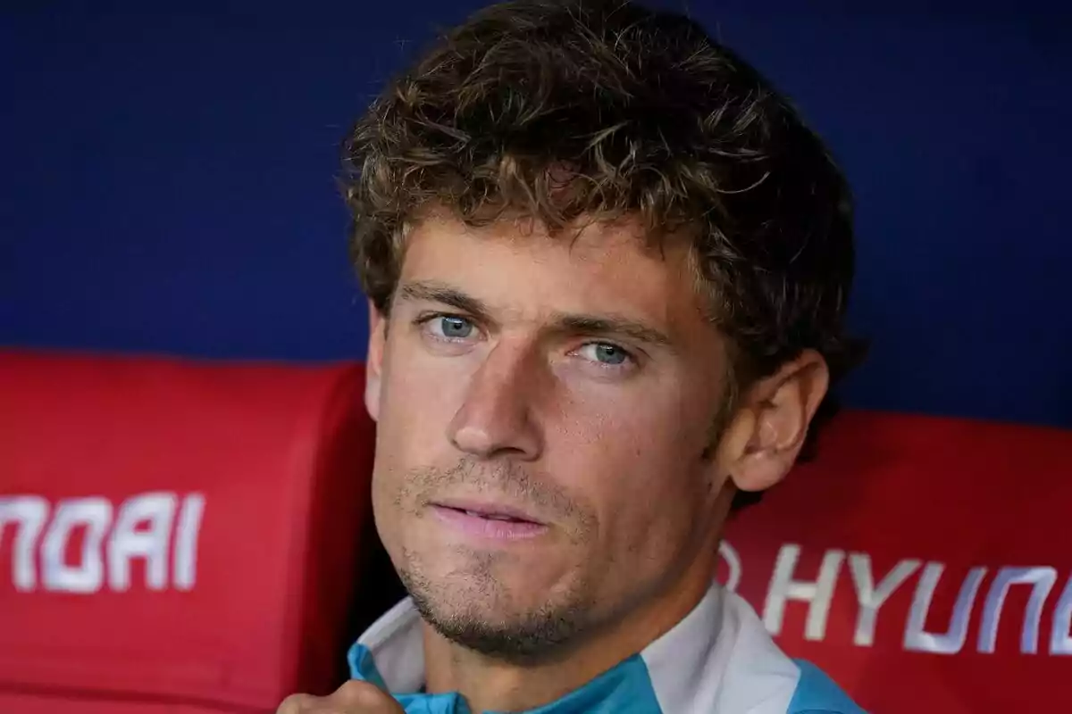 A man with curly hair and a serious expression is sitting in front of a blue and red background.