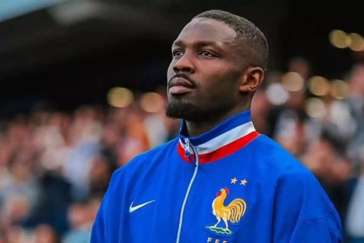 Un hombre con chaqueta azul de la selección de fútbol de Francia mira hacia arriba en un estadio lleno de espectadores.