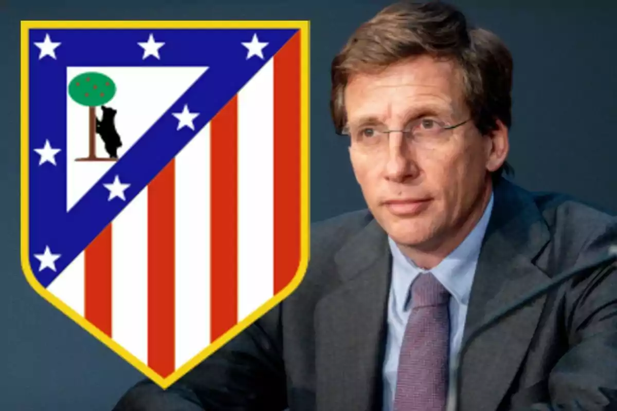 A man with glasses and a suit is sitting next to an Atlético de Madrid crest.