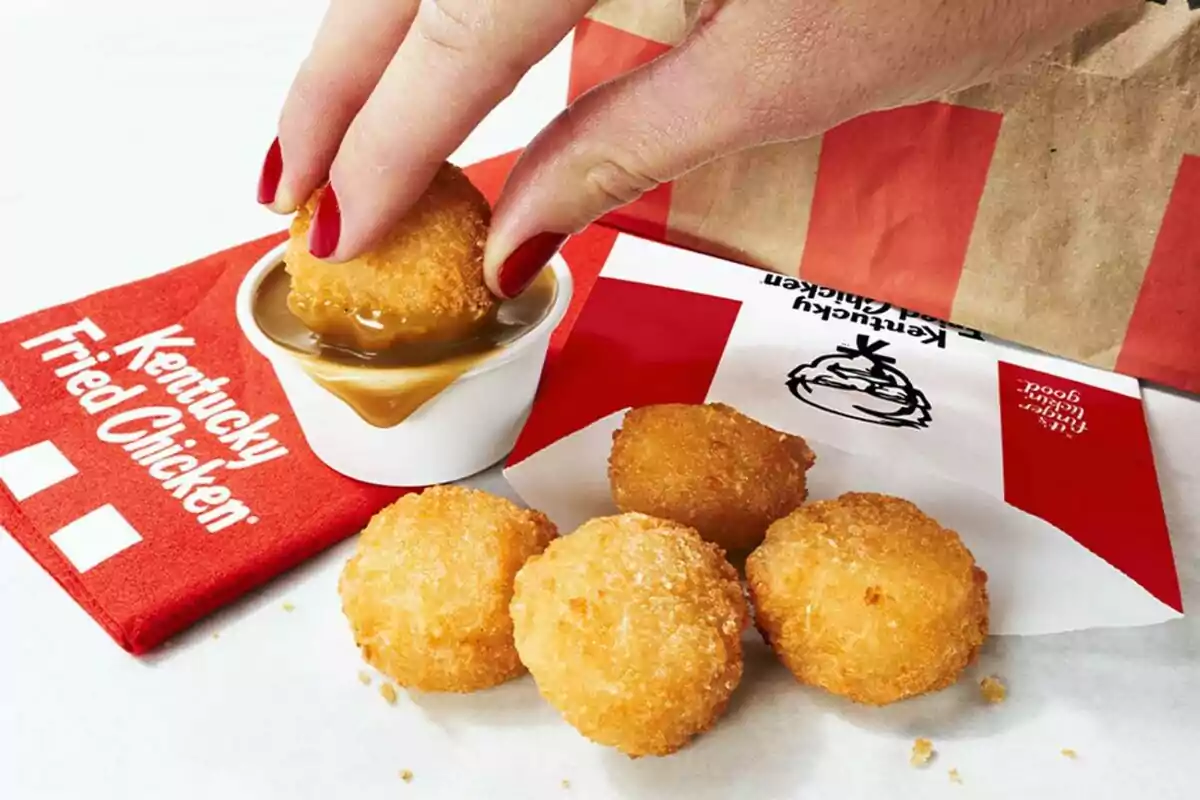 Una mano con uñas pintadas de rojo sumerge una bolita de patata empanizada en una salsa, rodeada de más bolitas sobre una servilleta roja de Kentucky Fried Chicken. Una mano con uñas pintadas de rojo sumerge una bolita de patata empanizada en una salsa, rodeada de más bolitas sobre una servilleta roja de Kentucky Fried Chicken.