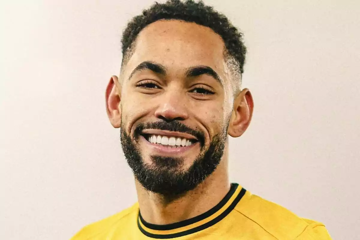 Un hombre sonriente con barba y cabello corto lleva una camiseta amarilla.