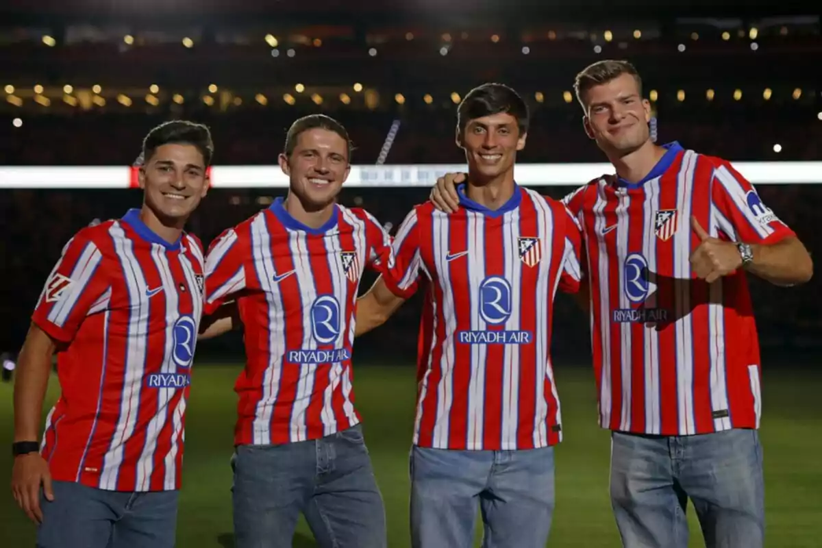 Cuatro hombres con camisetas del Atlético de Madrid posando juntos en un estadio.