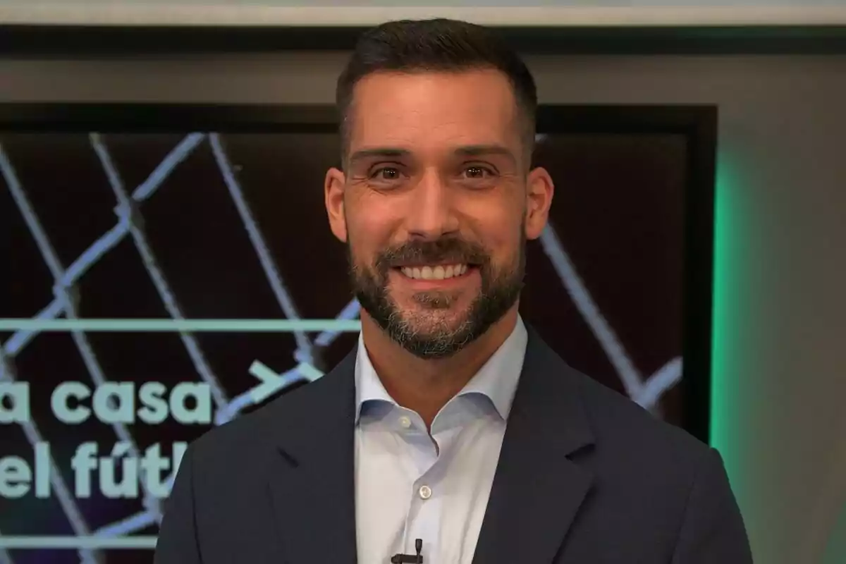 Un hombre con barba y traje sonríe frente a una pantalla con texto borroso.