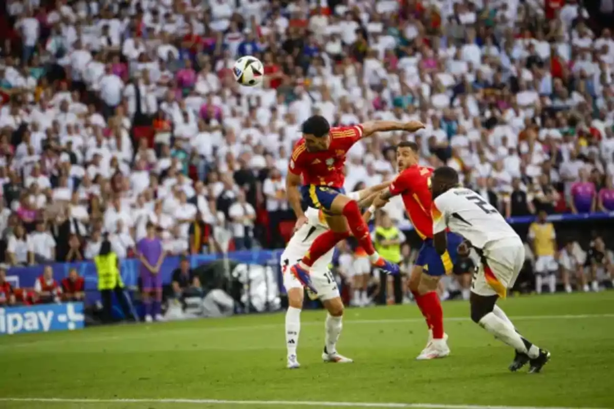 Jugadores de fútbol compiten por el balón en el aire durante un partido, con una multitud de espectadores en el fondo.