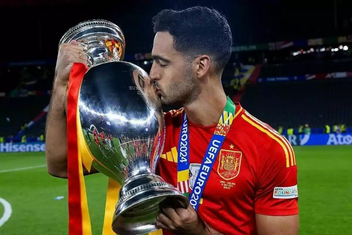 Jugador de fútbol con la camiseta de España besando un trofeo de la UEFA Euro 2024 en el campo de juego.