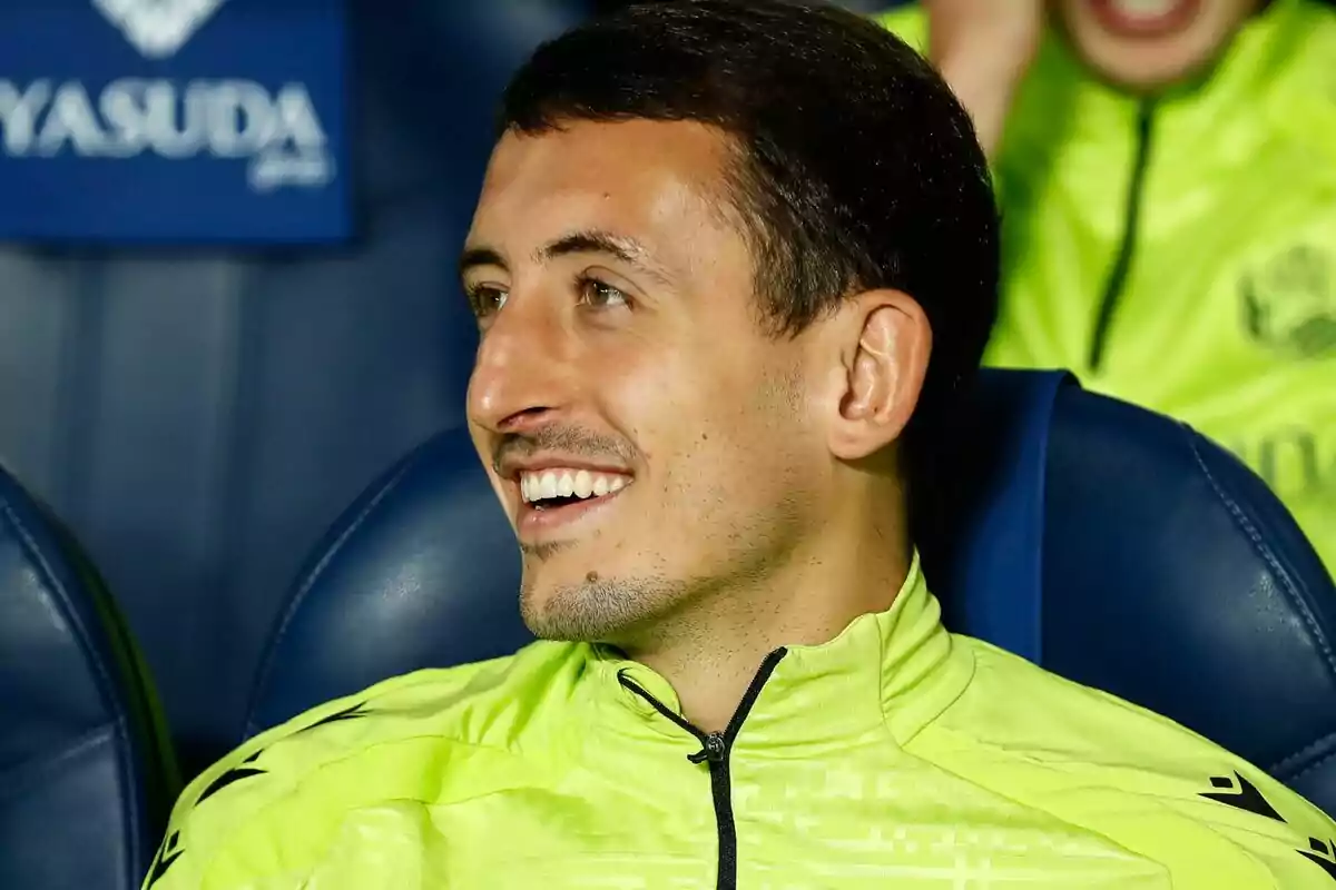 Un hombre sonriente con una chaqueta deportiva amarilla sentado en un asiento azul.