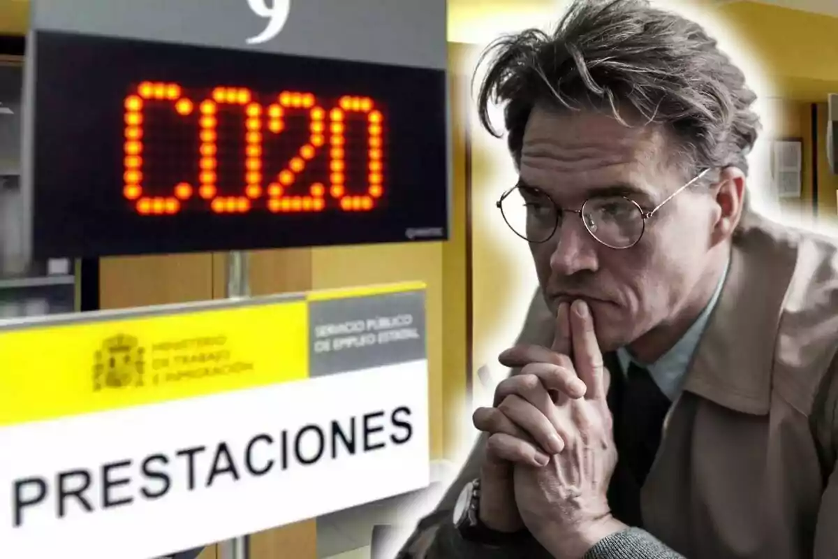 Un hombre con gafas y expresión pensativa está en una oficina de prestaciones, con un letrero electrónico que muestra el número C020.