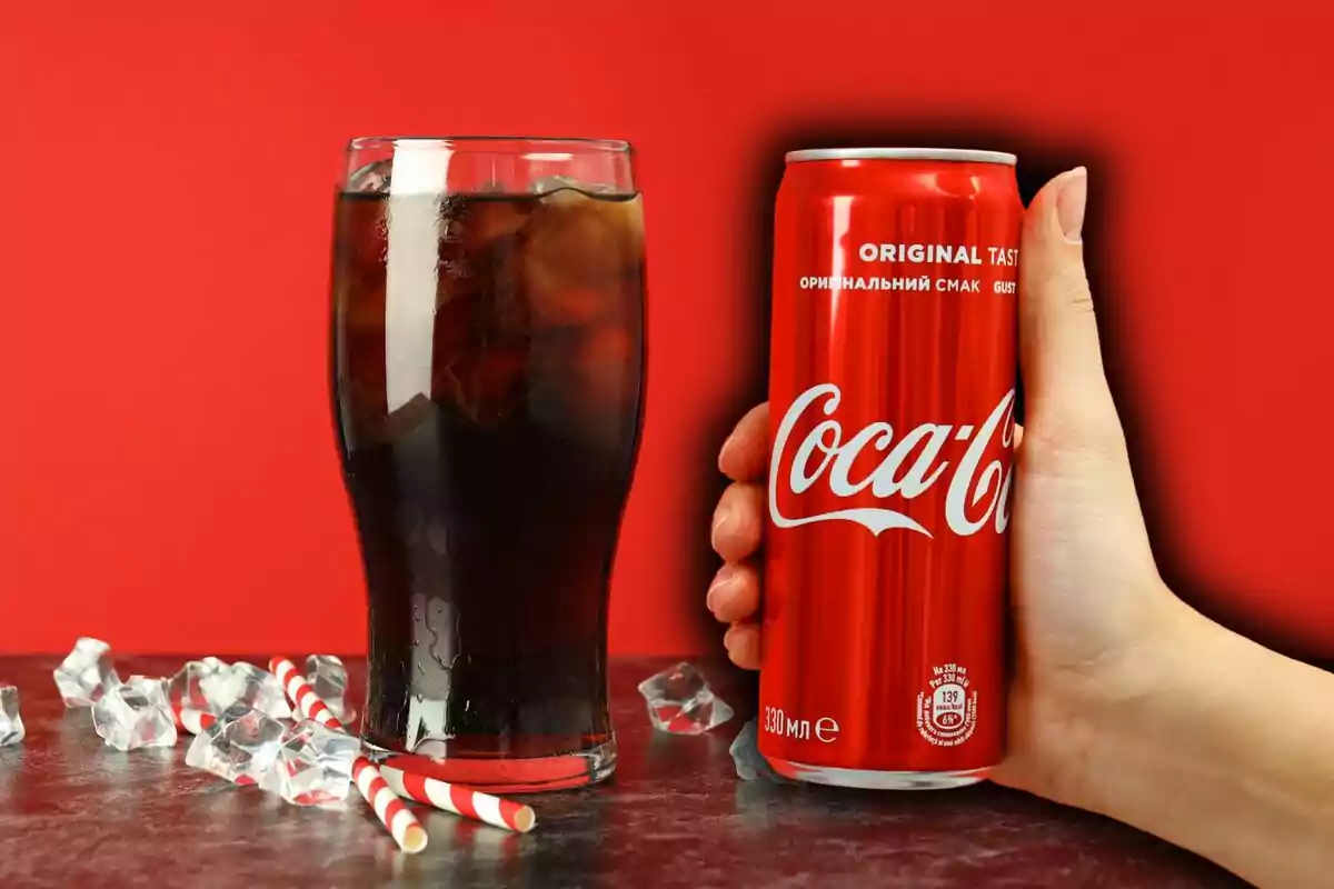 A hand holds a soda can next to a glass filled with a drink and ice on a surface with ice cubes and red and white straws, with a red background. A hand holds a soda can next to a glass filled with a drink and ice on a surface with ice cubes and red and white straws, with a red background.