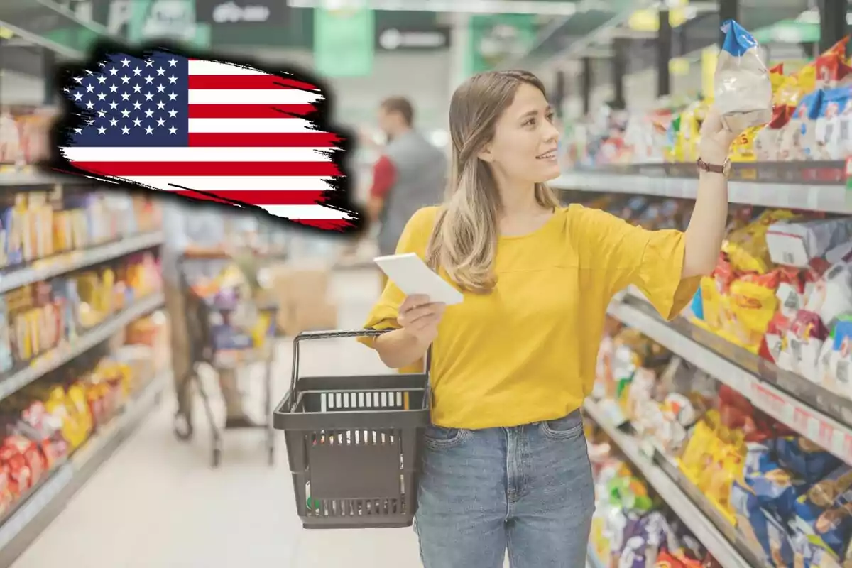 Una mujer con una blusa amarilla sostiene una canasta de compras y un teléfono mientras elige productos en un supermercado; en la esquina superior izquierda hay una bandera de Estados Unidos.