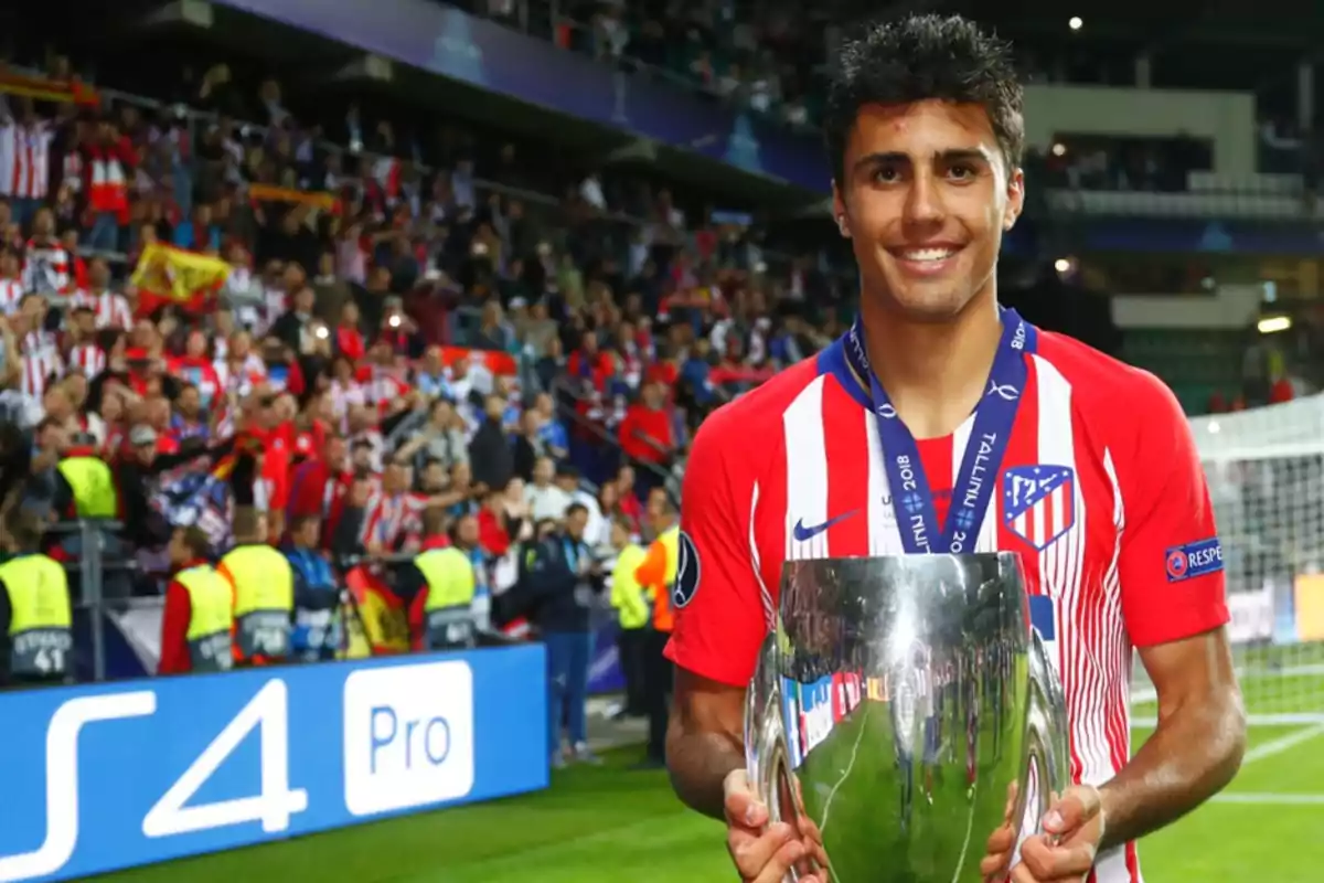 Un jugador de fútbol del Atlético de Madrid sostiene un trofeo en un estadio lleno de aficionados.