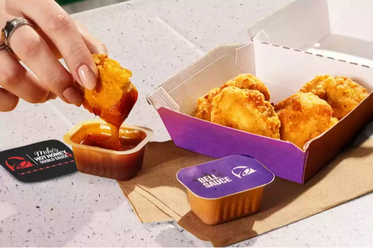 A hand dipping a breaded chicken piece into a sauce, with a box of nuggets and two sauce containers on a table. A hand dipping a breaded chicken piece into a sauce, with a box of nuggets and two sauce containers on a table.