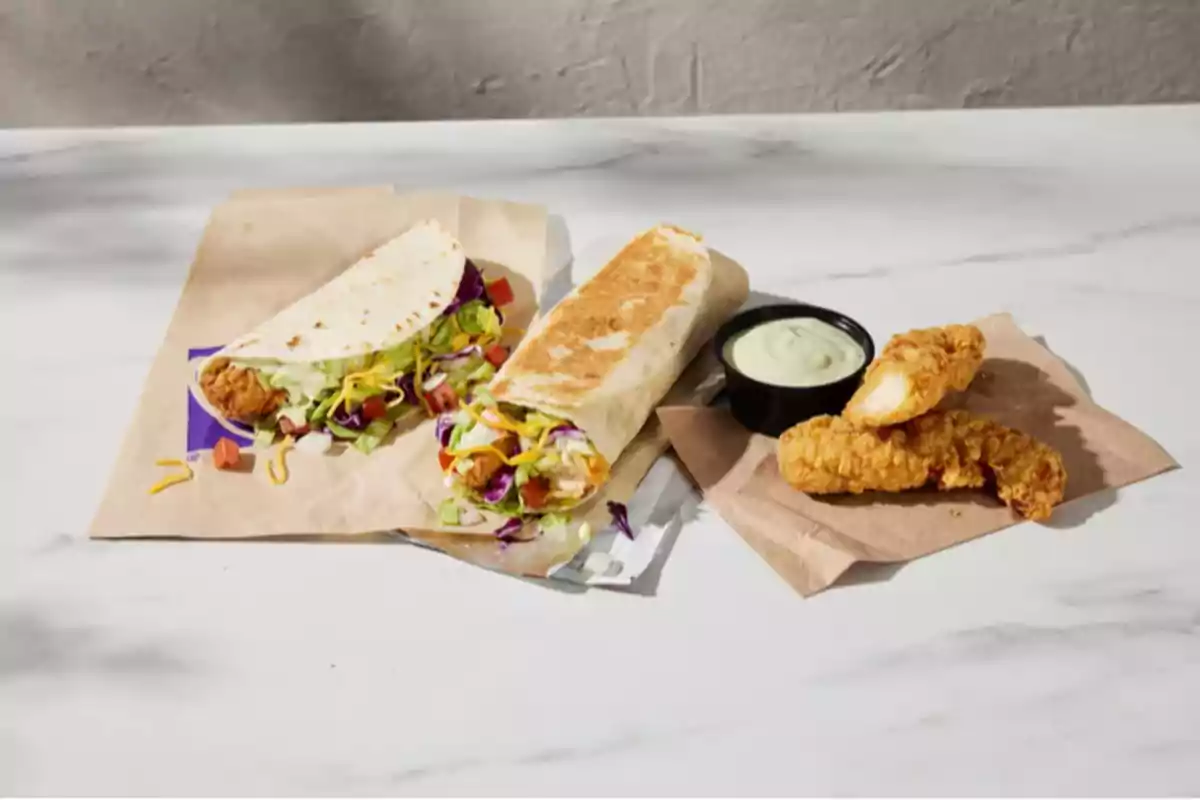 Three pieces of breaded chicken served with a creamy sauce, a taco and a burrito filled with chicken, lettuce, tomato, and cheese on a white table. Three pieces of breaded chicken served with a creamy sauce, a taco and a burrito filled with chicken, lettuce, tomato, and cheese on a white table.