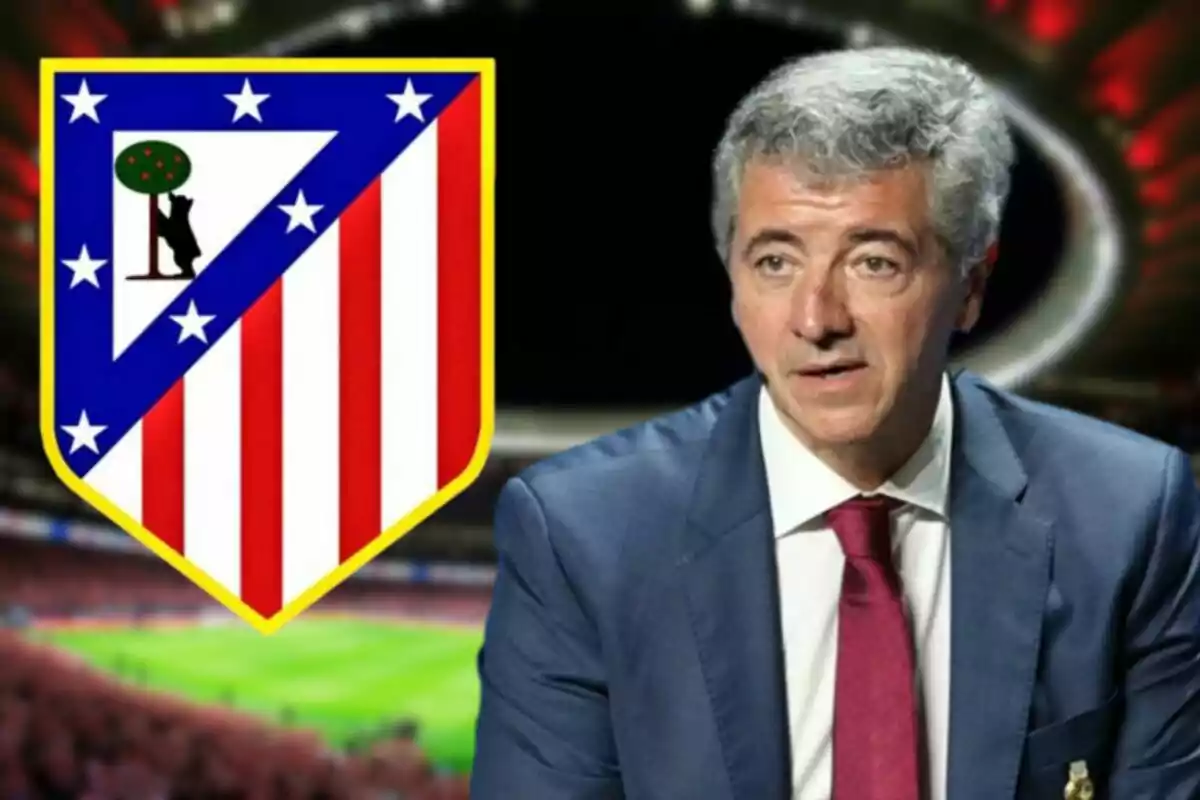 Un hombre con traje y corbata roja junto al escudo del Atlético de Madrid en un estadio.