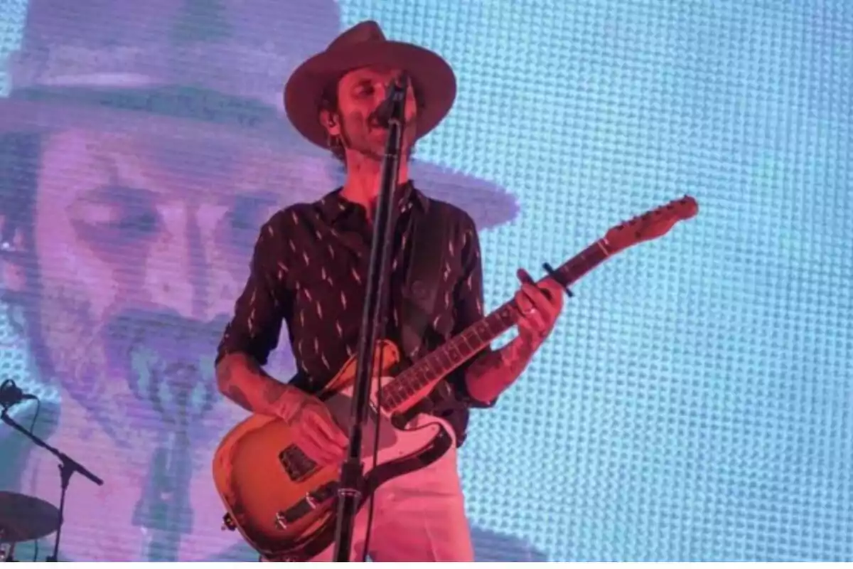 Un músico con sombrero toca la guitarra eléctrica en un escenario iluminado con una pantalla de fondo que muestra su imagen. Un músico con sombrero toca la guitarra eléctrica en un escenario iluminado con una pantalla de fondo que muestra su imagen.