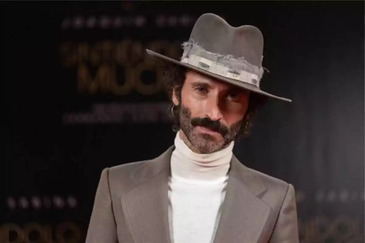 Un hombre con barba y bigote lleva un sombrero gris y un traje beige, posando frente a un fondo oscuro. Un hombre con barba y bigote lleva un sombrero gris y un traje beige, posando frente a un fondo oscuro.