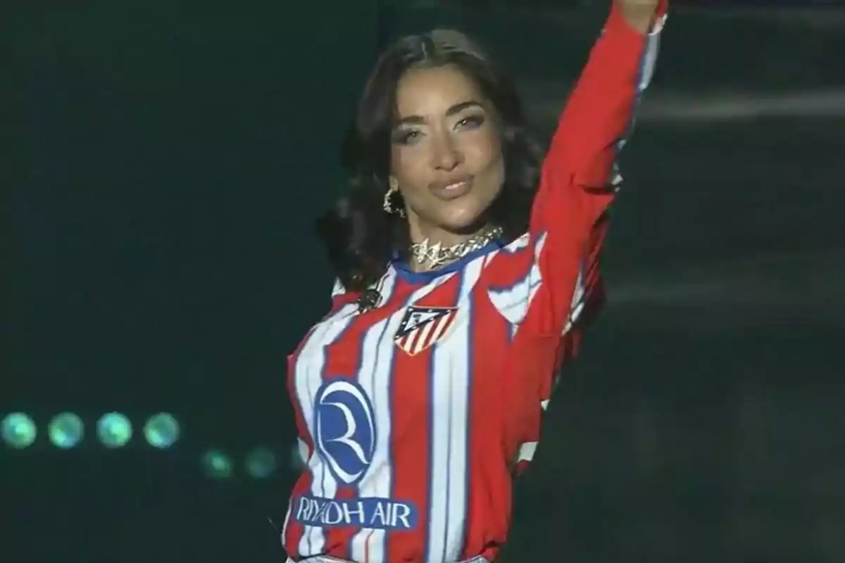 Una persona con una camiseta de rayas rojas y blancas con el logo de un equipo de fútbol levanta el brazo en un escenario iluminado. Una persona con una camiseta de rayas rojas y blancas con el logo de un equipo de fútbol levanta el brazo en un escenario iluminado.