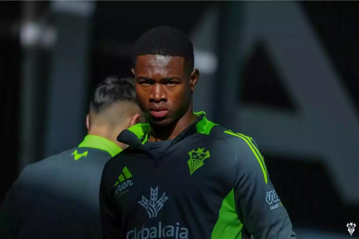 Un jugador de fútbol con uniforme gris y verde oscuro está de pie bajo una luz intensa, mirando hacia adelante con expresión seria, mientras otro jugador está de espaldas.