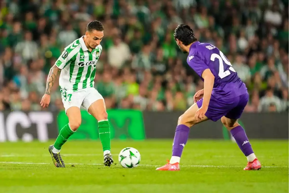Dos jugadores de fútbol en acción durante un partido, uno con uniforme verde y blanco y el otro con uniforme morado, en un estadio lleno de espectadores. Dos jugadores de fútbol en acción durante un partido, uno con uniforme verde y blanco y el otro con uniforme morado, en un estadio lleno de espectadores.