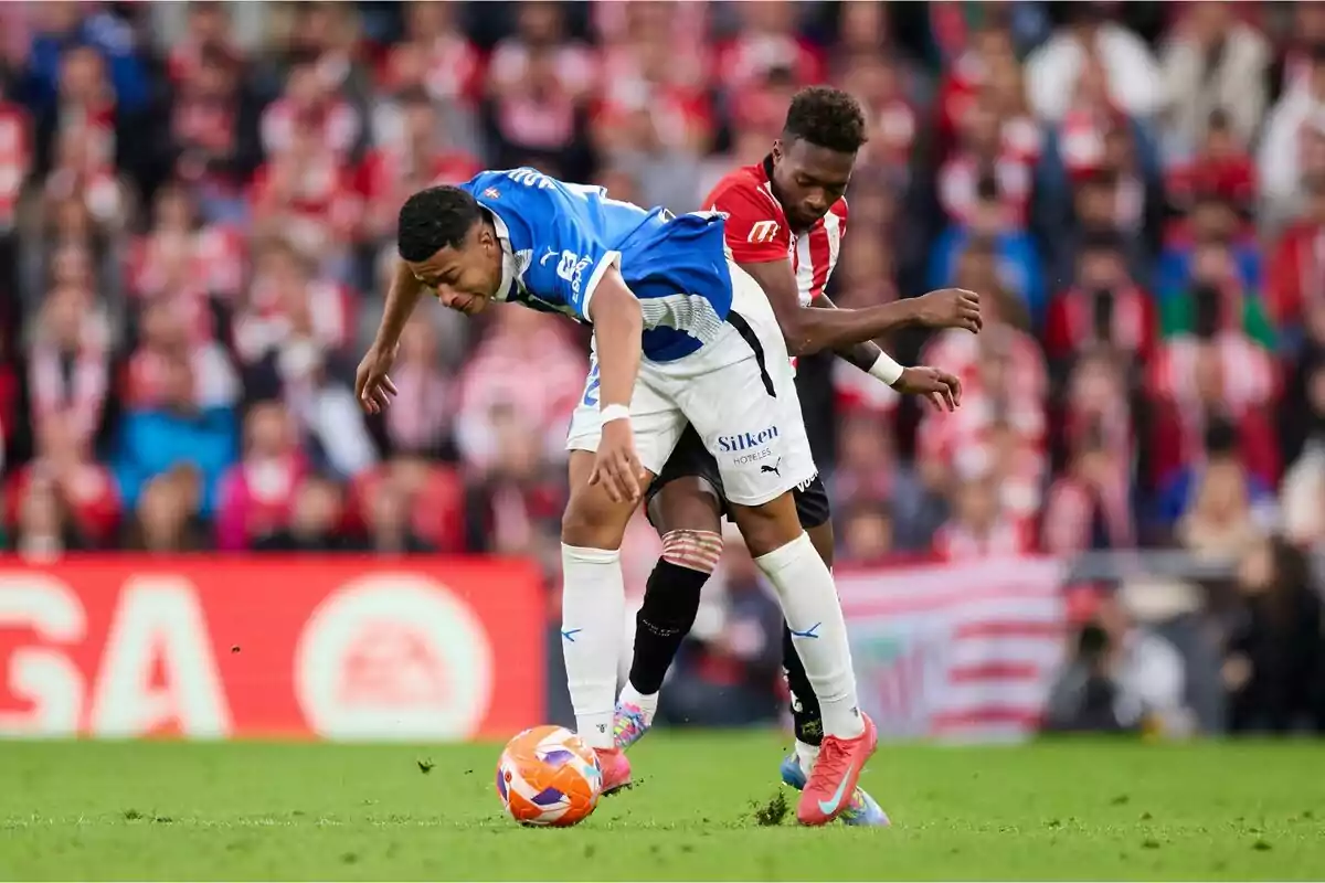 Dos jugadores de fútbol compiten por el balón en un partido, uno con uniforme azul y blanco y el otro con uniforme rojo y blanco, mientras el público observa desde las gradas. Dos jugadores de fútbol compiten por el balón en un partido, uno con uniforme azul y blanco y el otro con uniforme rojo y blanco, mientras el público observa desde las gradas.