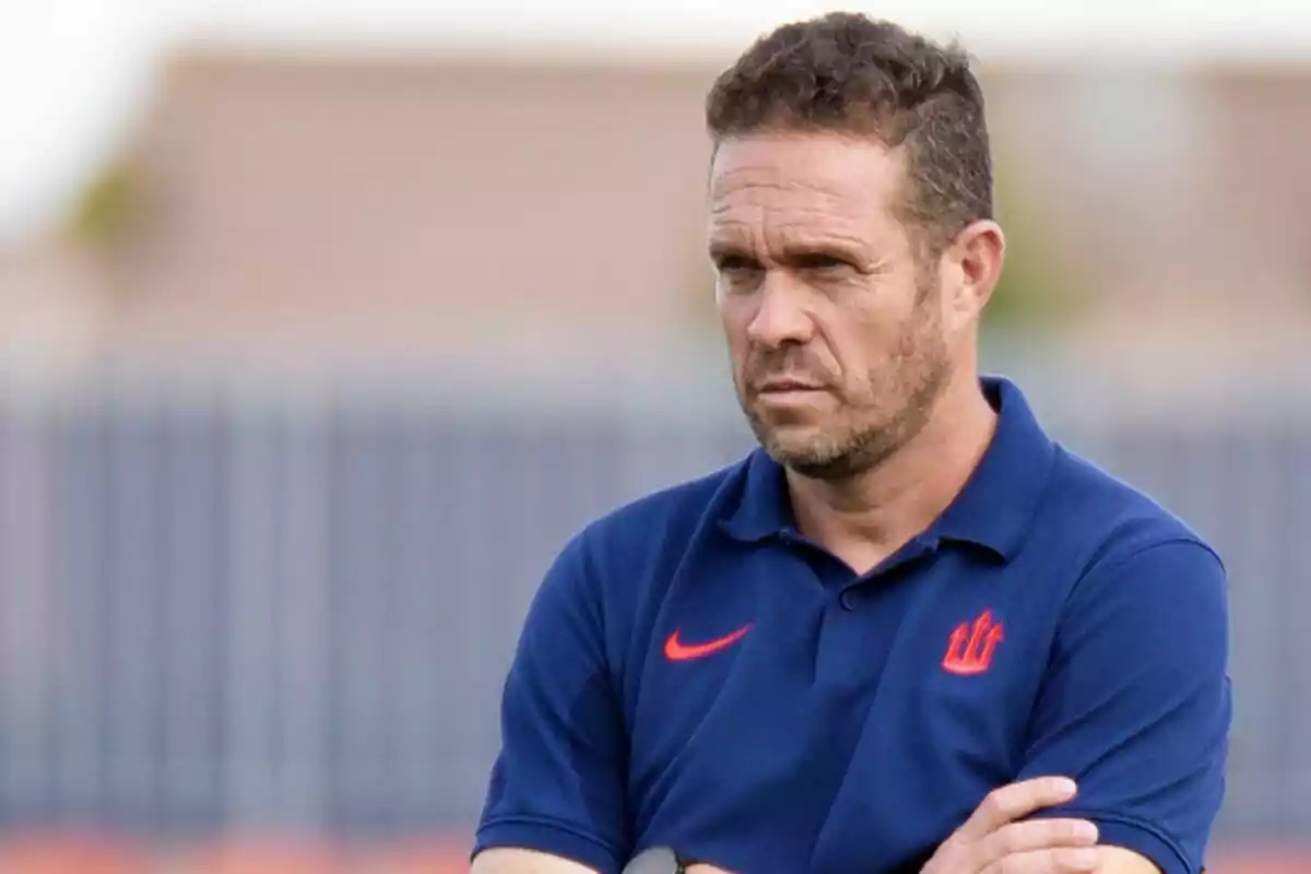 Un hombre con una camiseta azul y un logotipo rojo está de pie con los brazos cruzados, mirando hacia adelante con expresión seria. Un hombre con una camiseta azul y un logotipo rojo está de pie con los brazos cruzados, mirando hacia adelante con expresión seria.