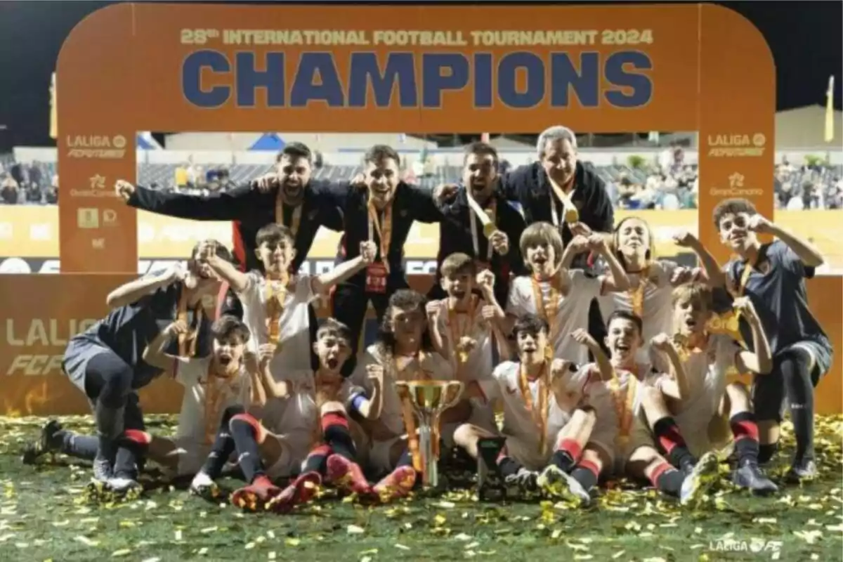 Un equipo de fútbol juvenil celebra su victoria en un torneo internacional, posando con medallas y un trofeo frente a un cartel que dice Un equipo de fútbol juvenil celebra su victoria en un torneo internacional, posando con medallas y un trofeo frente a un cartel que dice