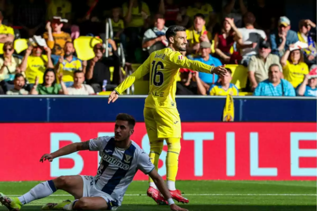 Un jugador de fútbol con uniforme amarillo señala mientras otro jugador con uniforme blanco y azul está sentado en el césped durante un partido, con espectadores en el fondo. Un jugador de fútbol con uniforme amarillo señala mientras otro jugador con uniforme blanco y azul está sentado en el césped durante un partido, con espectadores en el fondo.