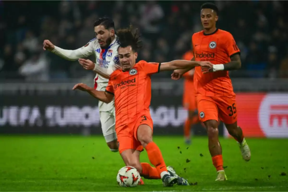 Jugadores de fútbol compiten por el balón en un partido, uno de ellos lleva uniforme naranja y el otro blanco.