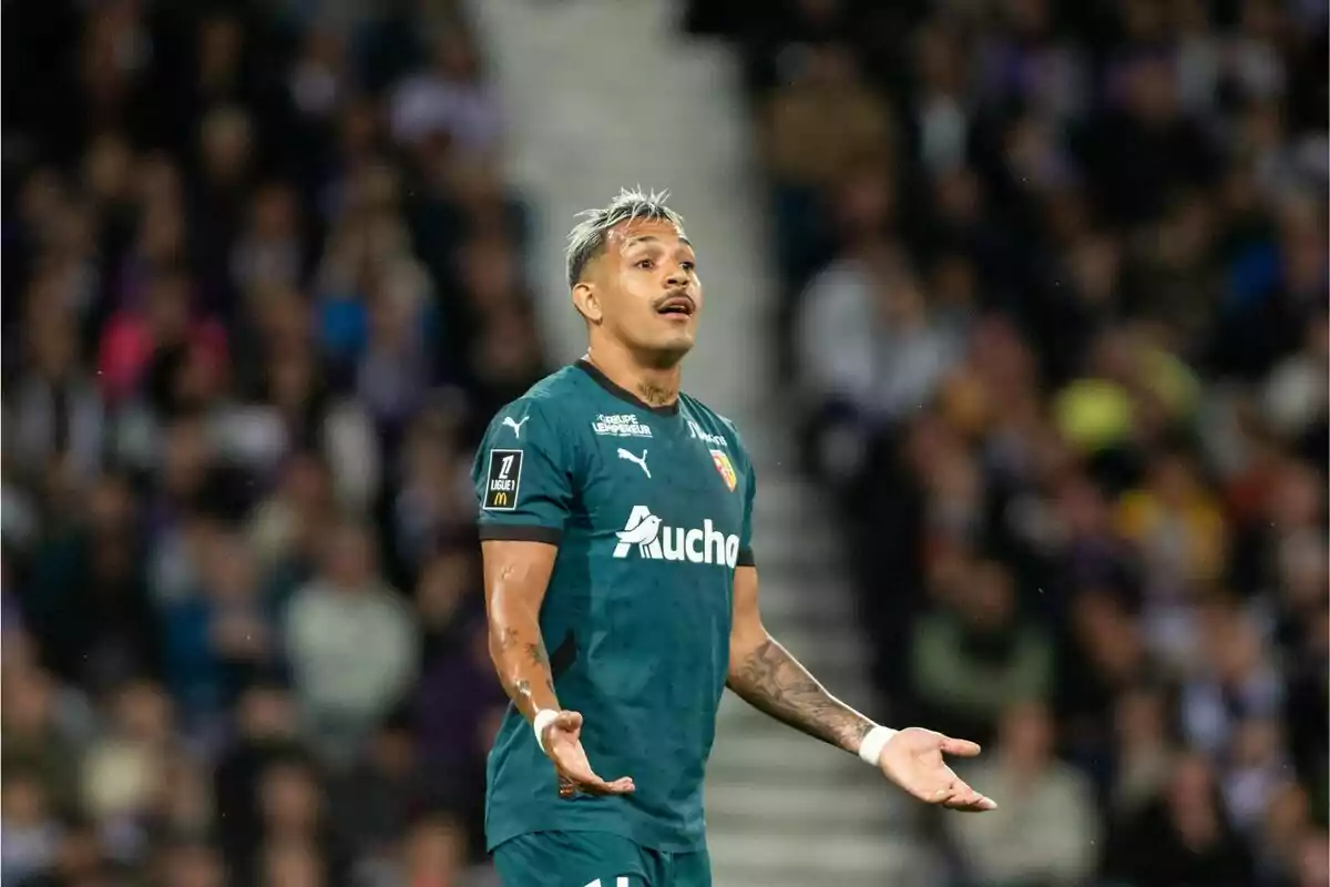 Un jugador de fútbol con uniforme verde oscuro gesticula con los brazos abiertos en un estadio lleno de espectadores.