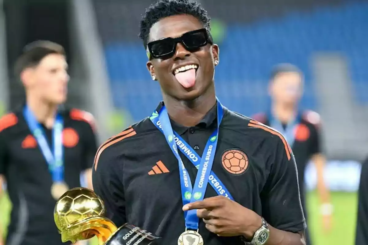 Jugador de fútbol con gafas de sol y medalla sonríe y saca la lengua mientras sostiene un trofeo dorado en un estadio Jugador de fútbol con gafas de sol y medalla sonríe y saca la lengua mientras sostiene un trofeo dorado en un estadio
