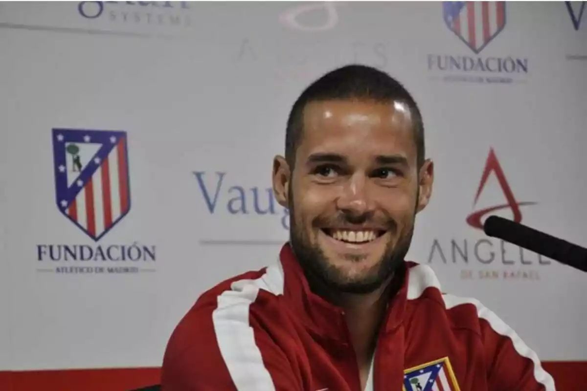 Hombre con chaqueta deportiva roja del Atlético de Madrid sonriendo durante una rueda de prensa con logotipos de patrocinadores en el fondo